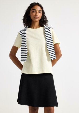 Kvinde med skulderlangt krøllet hår iført en bleggul kortærmet skjorte, sort nederdel og en stribet sweater lagt over skuldrene.