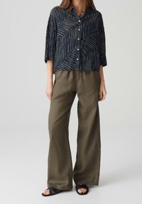 Camicia azzurra con bottoni e motivo astratto bianco, abbinata a pantaloni larghi in lino verde oliva, indossata con sandali neri con cinturini.