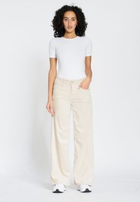 Beige wide-leg corduroy pants with a textured finish, paired with a fitted white t-shirt. Model stands in neutral background with white sneakers.