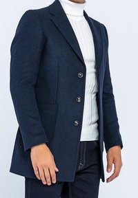 Mörkblå kavaj i strukturerad väv, med en enfaldig design, slagkrage och fem silverknappar, kombinerad med en vit polotröja.