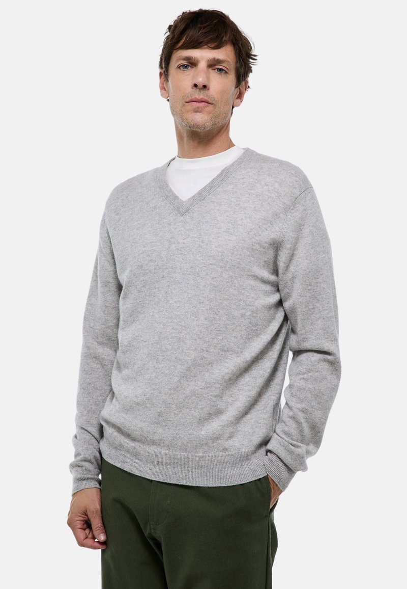 Homme portant un pull en V gris clair sur une chemise blanche avec un pantalon vert olive, se tenant avec une main dans sa poche.