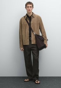 Brown suede button-up shirt over a black collared shirt, dark trousers, and flip-flops. A brown shoulder bag with a strap completes the look.