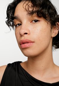 Gros plan sur un visage avec des cheveux noirs bouclés, une peau brun clair, des taches de rousseur et des lèvres roses douces, portant un haut en tissu noir sur fond blanc.