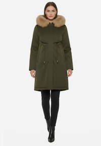 Parka verde oliva con colletto in pelliccia sintetica, chiusura con zip, vita regolabile con cordino e due tasche laterali. Texture liscia e design fino al ginocchio.