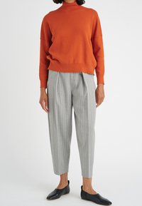 Kvinna som bär en orange långärmad tröja, ljusgrå randiga kortbyxor och svarta platta slip-on skor på vit bakgrund.