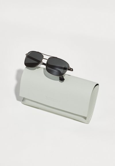 Ochelari de soare negri cu o formă rotundă și lentile gri, așezați pe un capac de culoare gri deschis, cu o textură moale, care prezintă un logo embosat "Burberry".