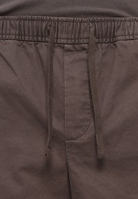 Gros plan sur un pantalon marron avec cordon de serrage, présentant une taille élastique et un détail de couture visible au niveau de la braguette.