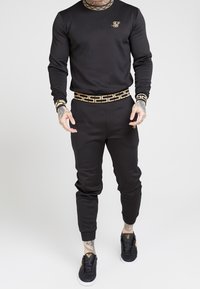 Svart tröja och joggers-set med guld accenter. Har en geometrisk design längs kragen och midjebandet, tillverkat av mjuk tyg.