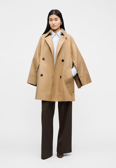 Νεαρή γυναίκα που φοράει μπεζ διπλόπατο παλτό, ανοιχτό μπλε πουκάμισο με ρίγες, σκούρα καφέ φαρδιά παντελόνια και καφέ παπούτσια, στέκεται μπροστά σε λευκό φόντο.