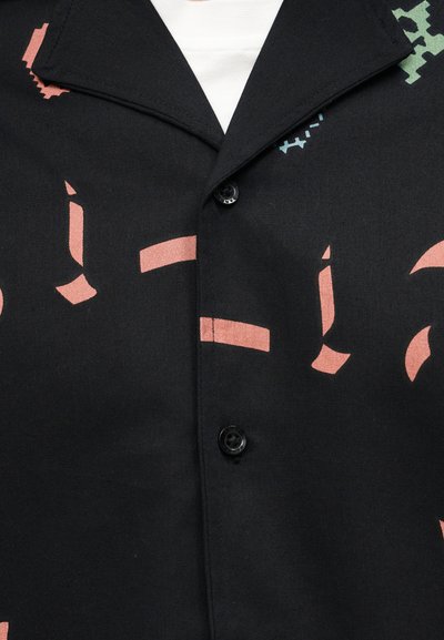 Μαύρο πουκάμισο με κουμπιά και φαρδιά γιακά, διακοσμημένο με αφηρημένα ροζ, πράσινα και μπλε σχήματα, φορεμένο πάνω από λευκό υποκάμισο.