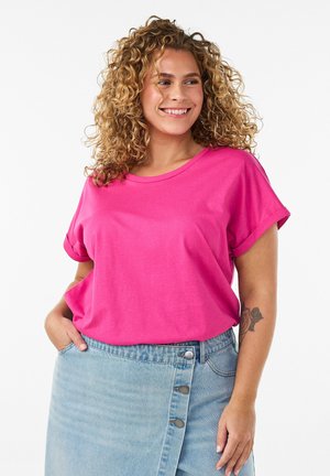 Mujer sonriente con cabello rizado que lleva una camiseta rosa brillante y una falda vaquera azul clara con botones, mano en el bolsillo, mostrando un tatuaje de rosa en el brazo.