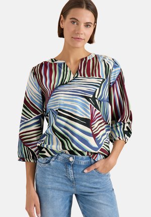 Frau mit kurzen braunen Haaren, die eine mehrfarbige gestreifte Bluse und hellblaue Jeans trägt, steht mit einer Hand in der Tasche.