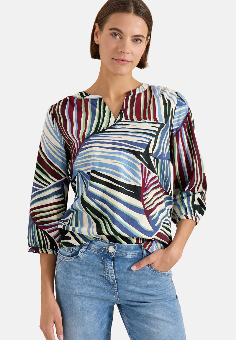 Femme aux cheveux courts bruns portant un chemisier rayé multicolore et un jean bleu clair, debout avec une main dans sa poche.