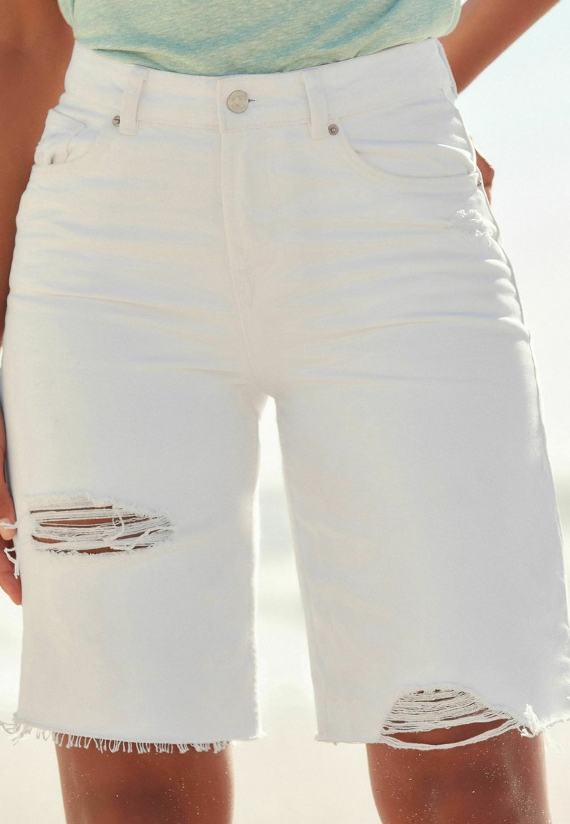 Personne portant un short en denim blanc déchiré avec des bords effilochés, se tenant à l'extérieur sous la lumière naturelle.