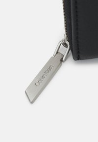 Czarna skórzana saszetka na suwak z metalowym uchwytem suwaka z wygrawerowanym logo "Calvin Klein", charakteryzująca się smukłym prostokątnym kształtem.