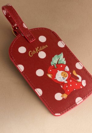 Punane pagasitempel valgete täppidega, Cath Kidstoni logo ja kass, kes kannab punast maasikamaski ning hoiab rohelist lehte.