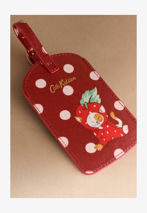 Punane pagasitempel valgete täppidega, Cath Kidstoni logo ja kass, kes kannab punast maasikamaski ning hoiab rohelist lehte.