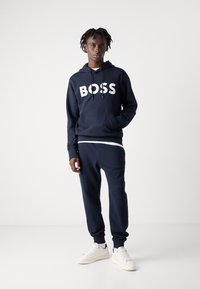 Sweatshirt azul-marinho com o logótipo branco "BOSS", combinado com calças de fato correspondentes. Apresenta um corte descontraído e punhos elásticos; usado com ténis brancos.