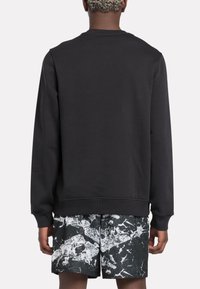 Persona con cabello rizado corto, vistiendo un sudadera negra lisa y pantalones cortos negros con un patrón abstracto blanco, de espaldas.