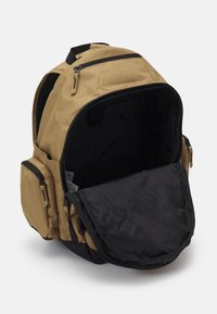 Mochila marrón claro con un interior negro. Cuenta con múltiples cremalleras, bolsillos laterales y un compartimento espacioso. Fabricada con tela duradera.