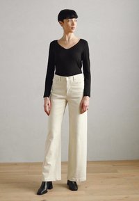 Black long-sleeve bodysuit paired with high-waisted, wide-leg beige jeans. Model wears black ankle boots, standing on a wooden floor.