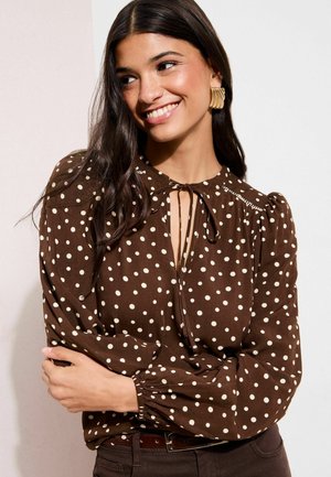 Braune Bluse mit weißen Punkten, Ausschnitt mit Schlitz, aufgeplusterten Ärmeln und elastischen Bündchen. Weicher Stoff, detaillierte Nahtführung entlang des Ausschnitts.