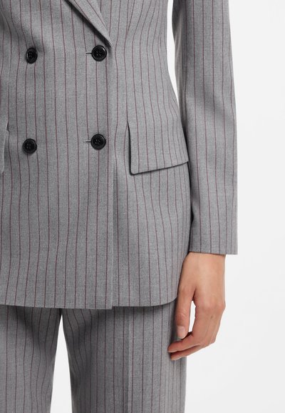 Costume gris croisé avec fines rayures verticales et boutons noirs, porté avec un pantalon rayé assorti, main détendue sur le côté.