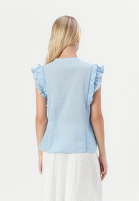 Top corto de manga corta en azul claro con detalles fruncidos. Hecho de una tela texturizada, con pequeños puntos tonales y un dobladillo curvado.