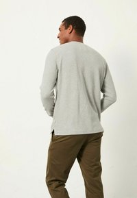 Graues Langarmshirt mit gerippter Textur und seitlichen Schlitzen, getragen mit olivgrünen Hosen. Einfacher Rundhalsausschnitt und dezentes Branding.