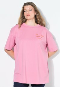 Rosa T-Shirt mit kurzen Ärmeln und lockerer Passform. Enthält orange gestickten Text auf der Brust. Weiches Baumwollmaterial.