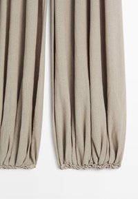 Beige linnen broek met geplooide elastische boorden, getoond vanaf halverwege het been naar beneden tegen een witte achtergrond.