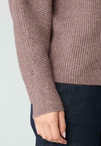 Różowy prążkowany sweter z miękkiej tkaniny, o dopasowanym kroju i długich rękawach, zestawiony z ciemnym denimu. Zbliżenie na rękaw.