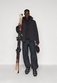 Skiausrüstung mit einer schwarzen Jacke und Hose, die ein Paar Skier hält. Die Skier sind braun mit schwarzen Bindungen und werden von einem schwarzen Helm ergänzt.