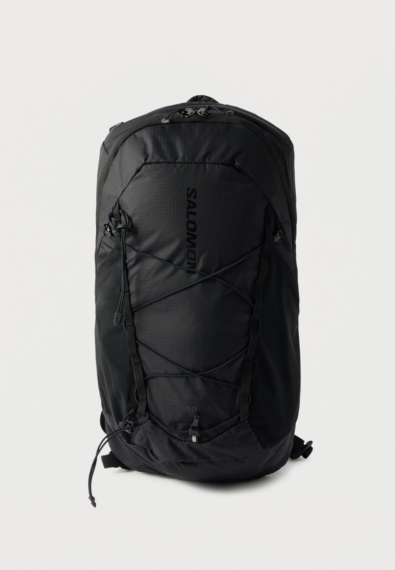 Μαύρο σακίδιο πλάτης Salomon XT 10 με μπροστινούς ελαστικούς ιμάντες, πλαϊνές τσέπες από δίχτυ και επάνω φερμουάρ σε λευκό φόντο.