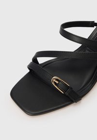 Sandale noire à bout carré avec de fines lanières en cuir et une petite boucle dorée sur la lanière avant sur un fond clair.