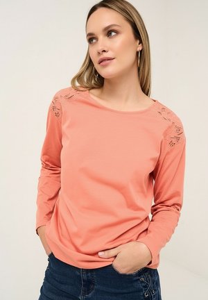 Frau trägt ein korallenfarbenes Langarmshirt mit Spitzenverzierung auf den Schultern, posiert mit den Händen in den Taschen und schaut leicht zur Seite.