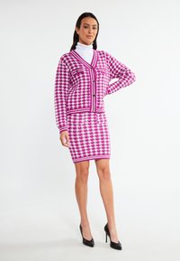 Rosa och vit houndstooth-stickad cardigan med V-ringning, knappar och två framfickor, matchad med en figurnära kjol. Svarta klackar.