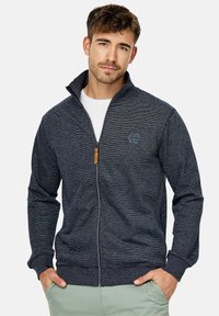 Mørkeblå zip-up jakke i struktureret stof, med høj krave, ribbede manchetter og et logo-patch på brystet.