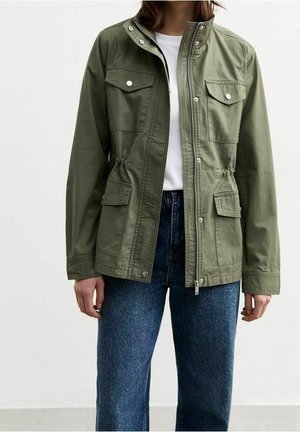Kvinde iført en olivengrøn jakke med knapper og lynlås, med bryst- og hoftelommer, kombineret med blå jeans og en hvid skjorte under.