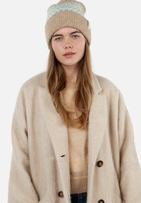 Young woman with long light brown hair wearing a beige knit beanie, beige sweater, and oversized beige wool coat against white background.