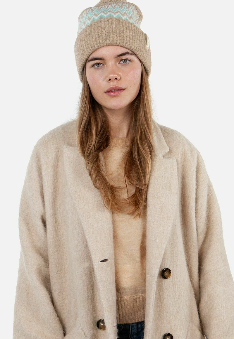 Young woman with long light brown hair wearing a beige knit beanie, beige sweater, and oversized beige wool coat against white background.