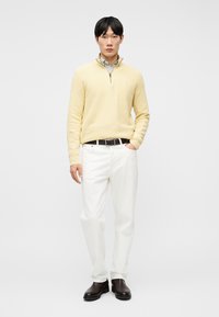 Mladi moški stoji z roko v žepu, oblečen v bledo rumen pulover z zadrgo prek modre srajce s črtami, bele hlače, črni pas in temne usnjene škornje.