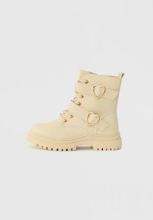 Beige ankle boot with chunky sole, featuring gold heart buckles and laces. Made of smooth material, with two adjustable straps.