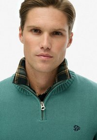 Miętowy sweter dziany z zamkiem przy kołnierzu, noszony na czarnej koszuli w kratę. Posiada ściągacze na rękawach i logo na piersi.