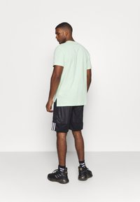 Camiseta de manga corta verde claro con aberturas laterales, combinada con pantalones cortos de malla negros con rayas blancas y zapatillas deportivas negras.