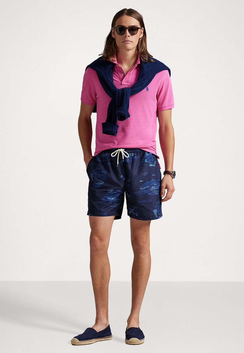 Chemise polo rose, shorts de bain à motif bleu marine, pull bleu marine noué autour du cou, lunettes de soleil, et espadrilles aux pieds.