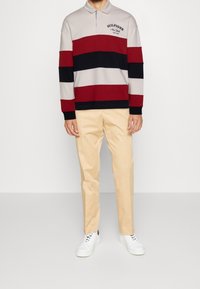 Polo a maniche lunghe con righe orizzontali in grigio, bordeaux e blu navy; logo ricamato sul colletto. Abbinato a pantaloni beige e sneakers bianche.
