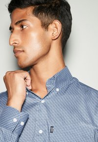 Camicia blu a fantasia con colletto button-down, dotata di bottoni bianchi e un piccolo logo sulla parte centrale, tessuto dalla texture liscia.