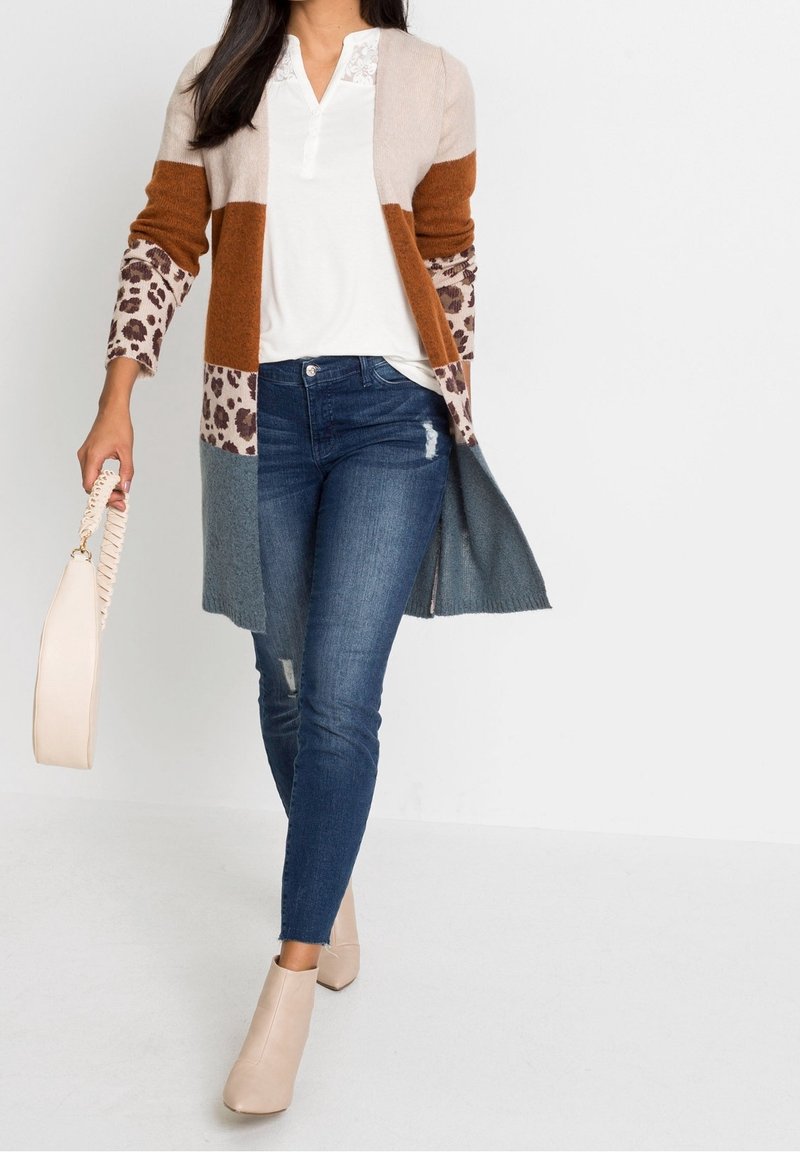 Stickad cardigan med färgblock i beige, brunt och leopardmönster; kombinerad med en vit topp och blå skinny jeans; håller en ljus väska.