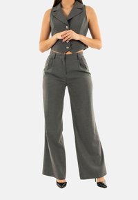 Panciotto grigio su misura con colletto a punte e tre bottoni, abbinato a pantaloni grigi a gamba larga. Il materiale ha una texture sottile. TACCHI NERI.